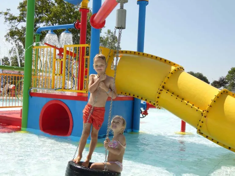 Twee kinderen in het ondiepe buitenbad met waterspeeltoestel en glijbaan op Camping Zonneweelde